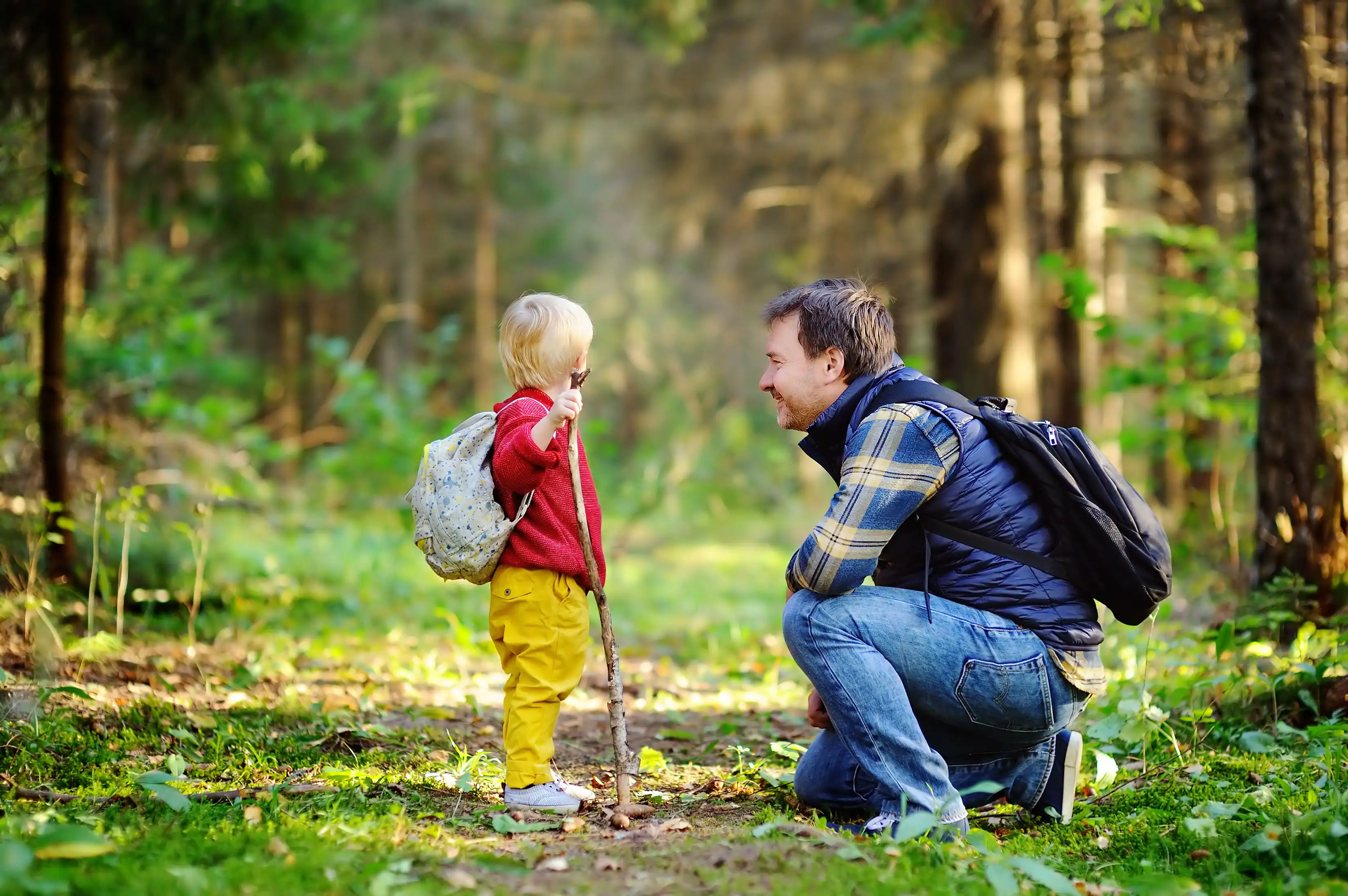 father crouching down to sons level and smiling, little boy looking toward the woodland trail holding a large stick in his right hand
